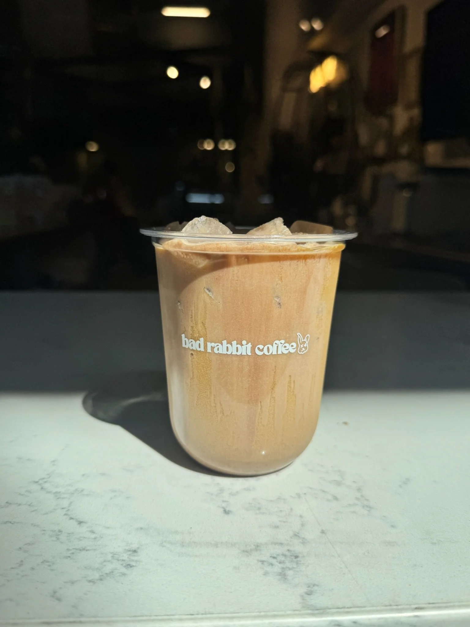 A clear plastic cup filled with iced coffee, with the words 'bad rabbit coffee' and a bunny logo printed on it, sitting on a white surface.