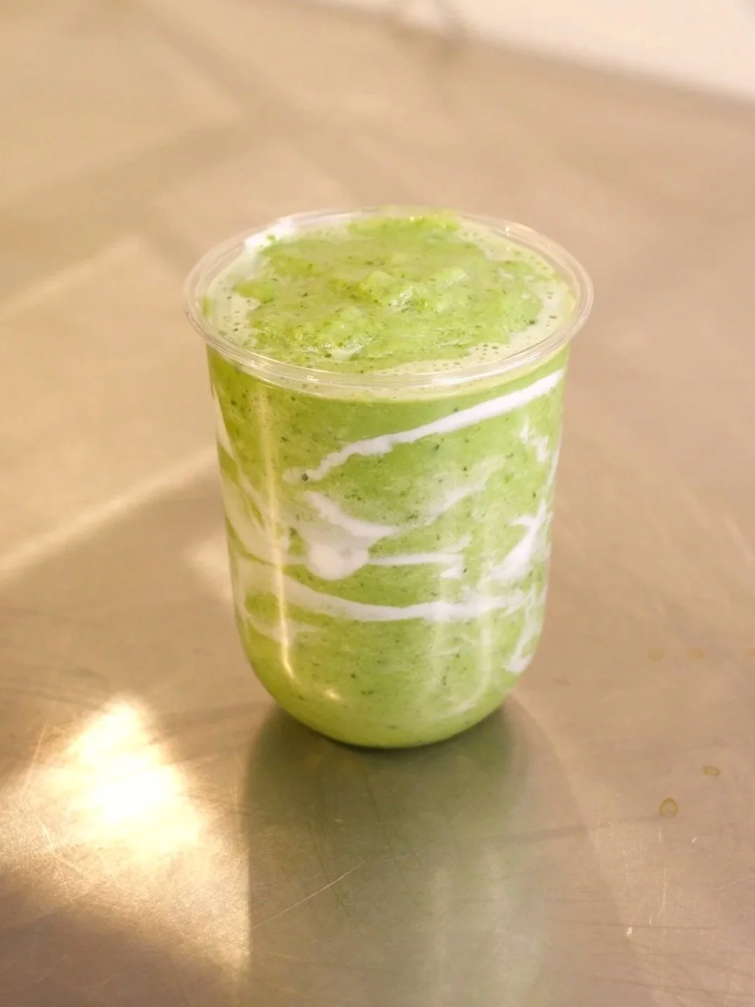 Green smoothie with white swirls in a clear plastic cup on a metallic surface.