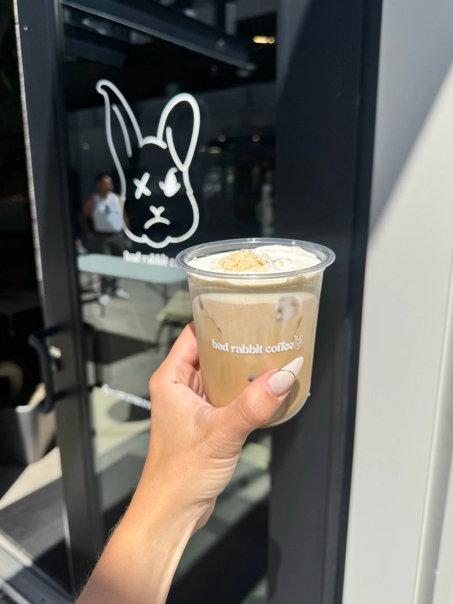 A hand holding a plastic cup of iced mocha coffee at Bad Rabbit Coffee with a black shelf in the background featuring a white outline of a bunny's face with one eye crossed out and the other eye as an 'O' shape.