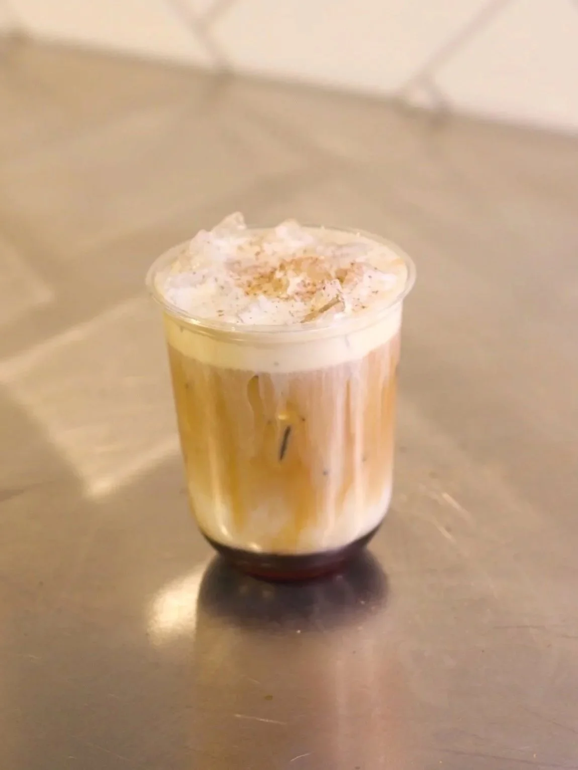 A caramel-colored coffee drink topped with whipped cream and a sprinkle of cinnamon in a clear plastic cup on a metallic surface.