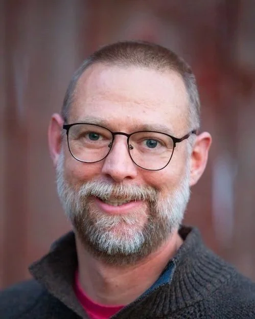 Close-up of a smiling middle-aged man with glasses and a beard, standing in front of a red brick wall.