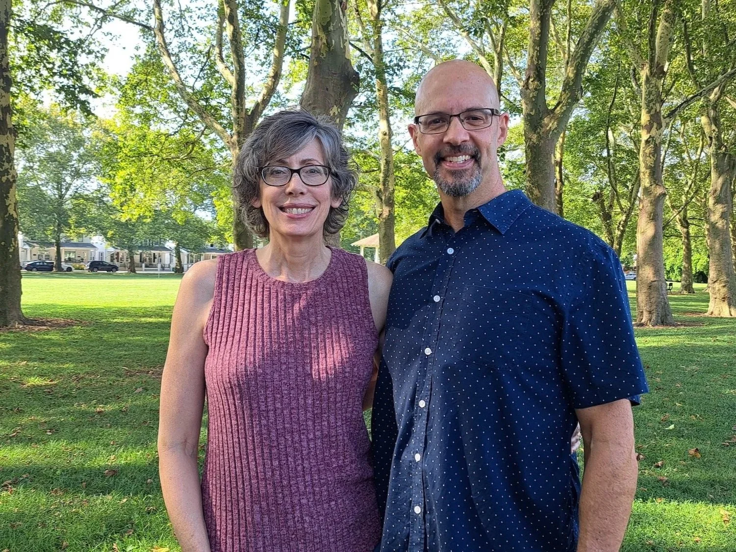 A man and a woman standing in a park with trees and houses in the background. Both are smiling and wearing glasses.