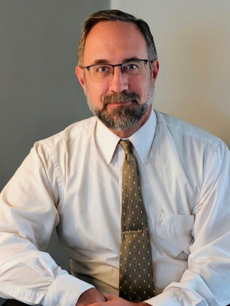 Close-up of a smiling middle-aged man with glasses and a salt-and-pepper beard, sitting on a gray patterned chair against a plain light-colored wall.