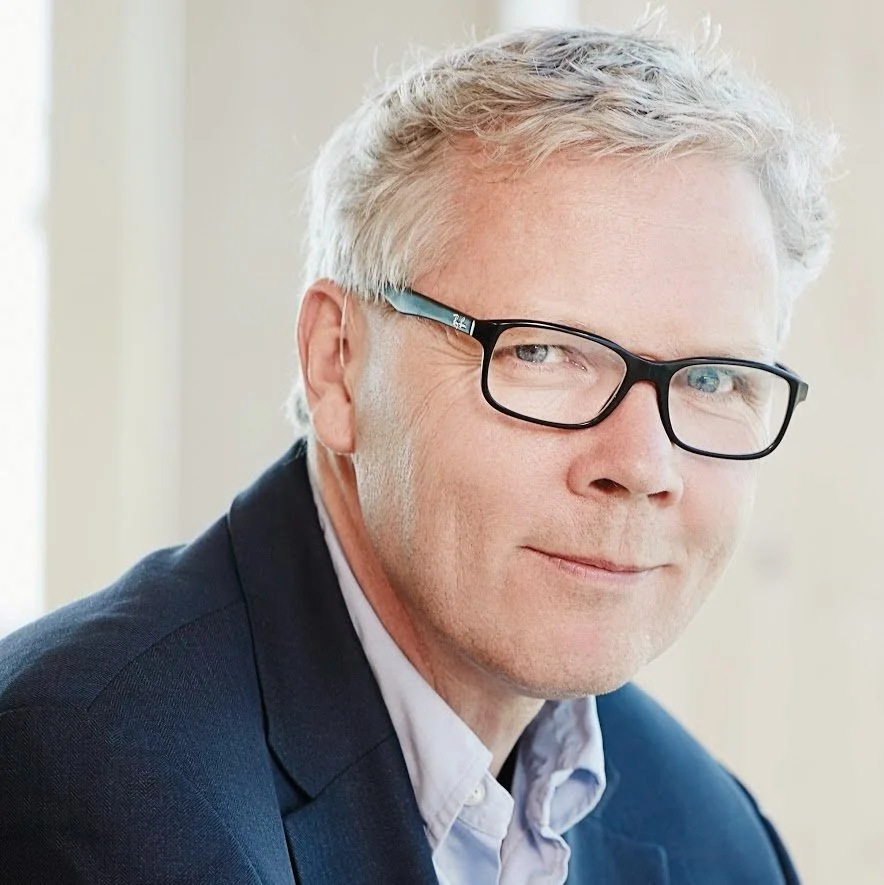 A middle-aged man with gray hair and glasses, wearing a navy blazer and light shirt, looking at the camera with a slight smile.