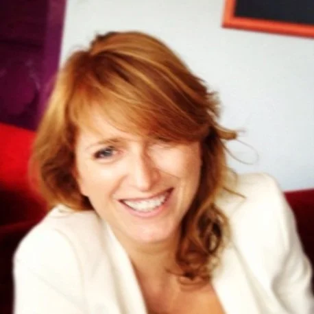 A smiling woman with red hair, wearing a white jacket, in an indoor setting with purple and white background elements.