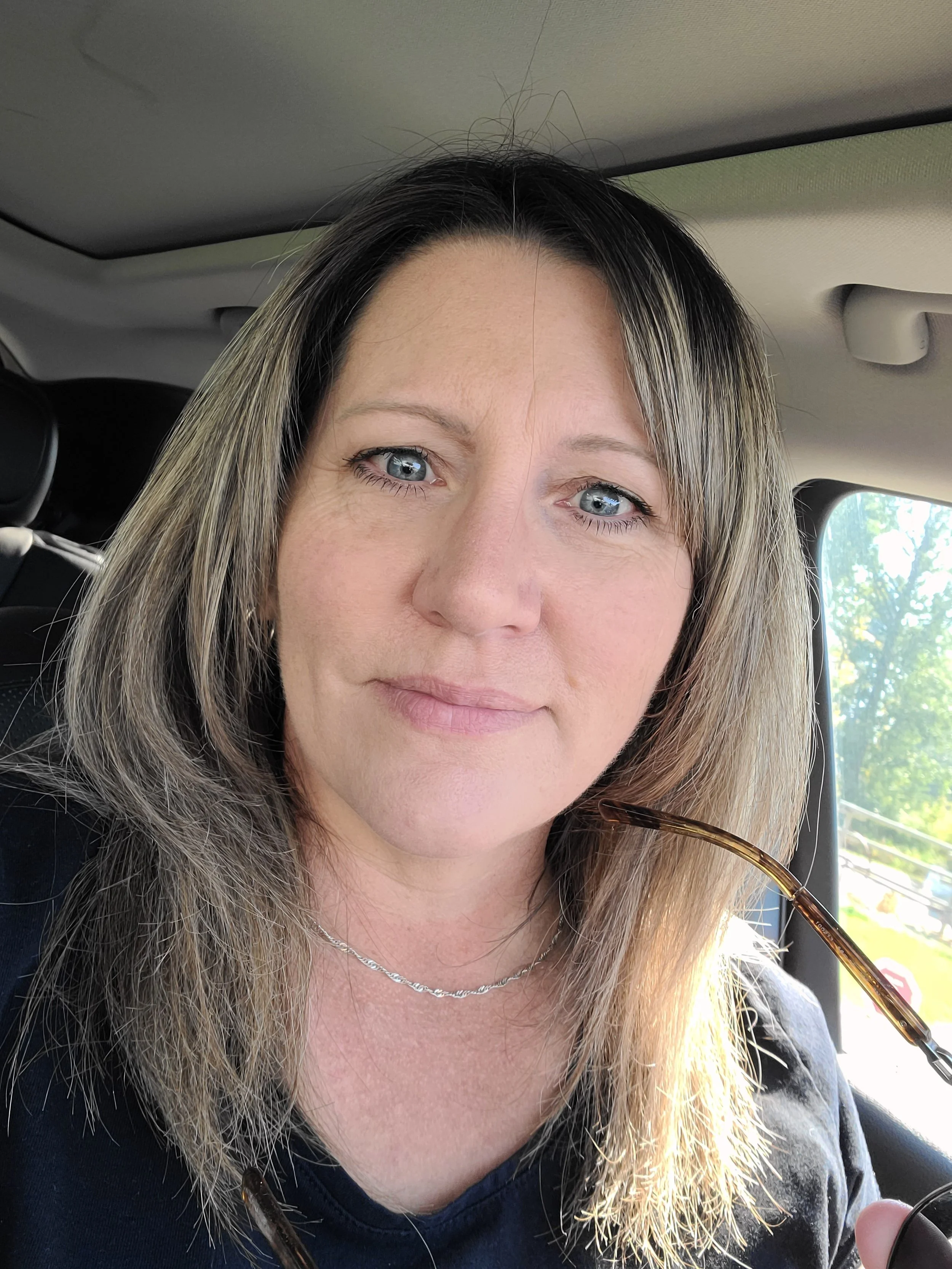 A woman with shoulder-length light brown hair and blue eyes sitting in a car, holding sunglasses, with trees visible outside the window.