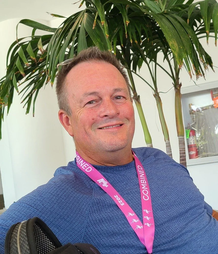 A man smiling in front of a green plant, wearing a blue athletic shirt and a pink lanyard that reads '2023 CO-MIXED' with small flamingo icons.
