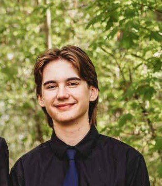 A young man with brown hair, smiling, wearing a black shirt and blue tie, outdoors with green leafy trees in the background.
