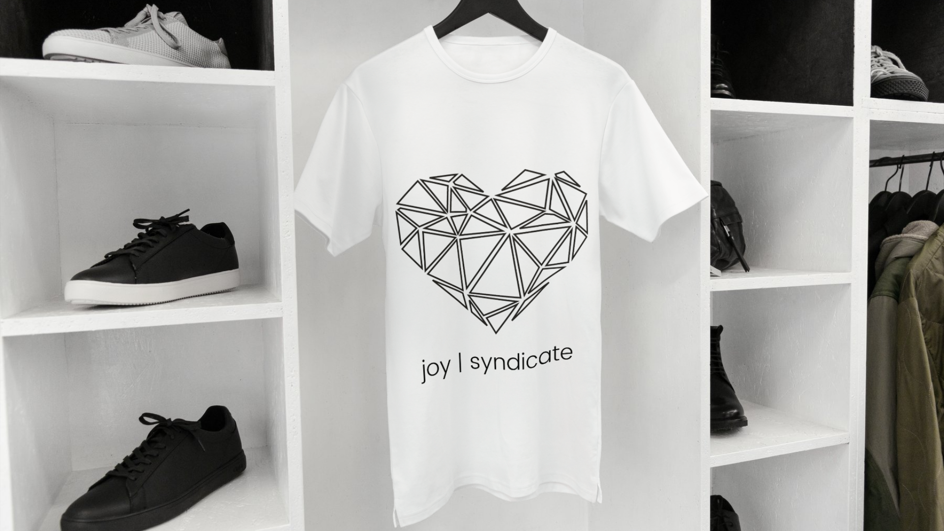 A white T-shirt with a geometric heart design and the text 'joy | syndicate' hanging on a black hanger in a closet. Surrounding it are shelves with black shoes and other clothing items.