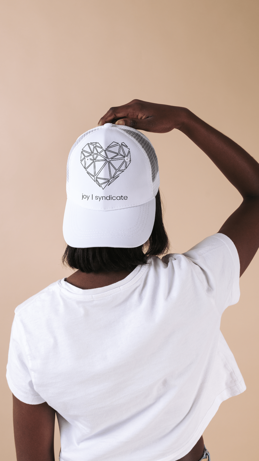 Person wearing a white T-shirt and a white baseball cap with a geometric heart design and the text 'joy | syndicate' in front of a beige background.