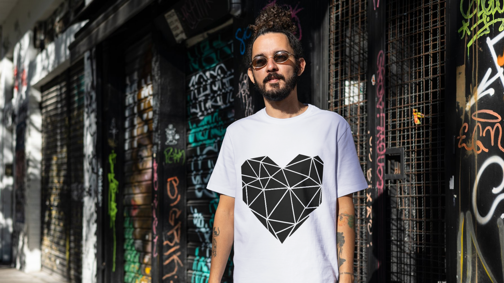 A man wearing sunglasses and a white t-shirt with a black geometric heart design standing in front of a graffiti-covered wall.