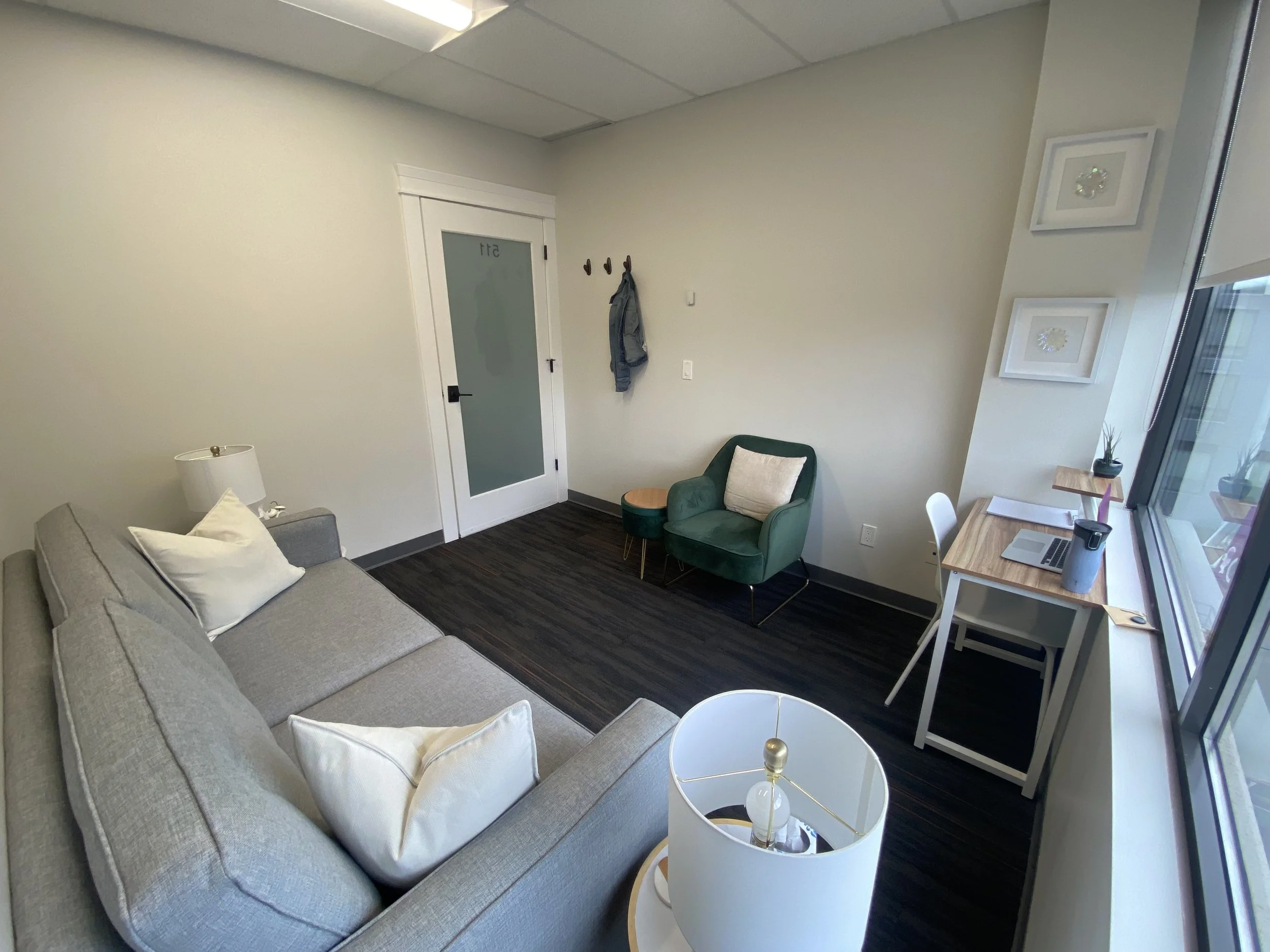 A modern therapy office with a gray sofa, white pillows, a small green armchair with a white pillow, a wooden desk with a laptop, water bottle, and plant, a white side table with a lamp, dark flooring, and a large window.