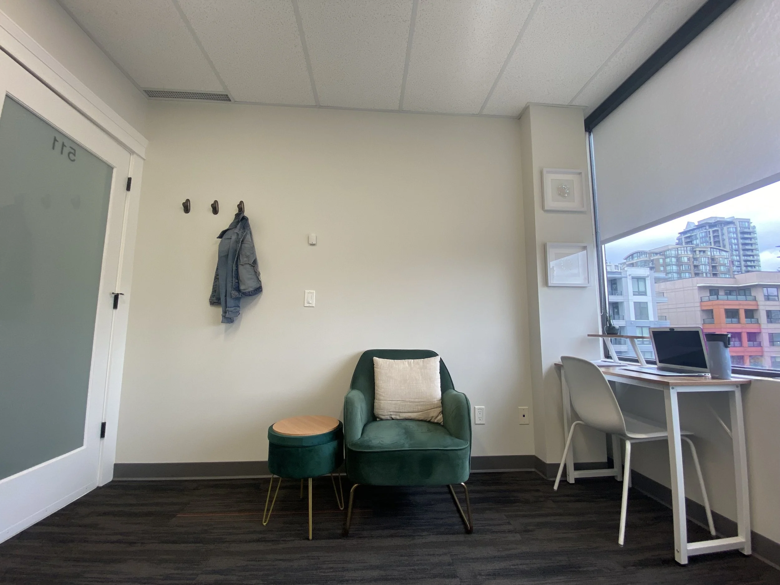 An office with a green armchair, a small matching storage ottoman, a white desk with a chair, a laptop, and a water bottle, near a large window with city buildings outside, and four framed pictures on the wall.
