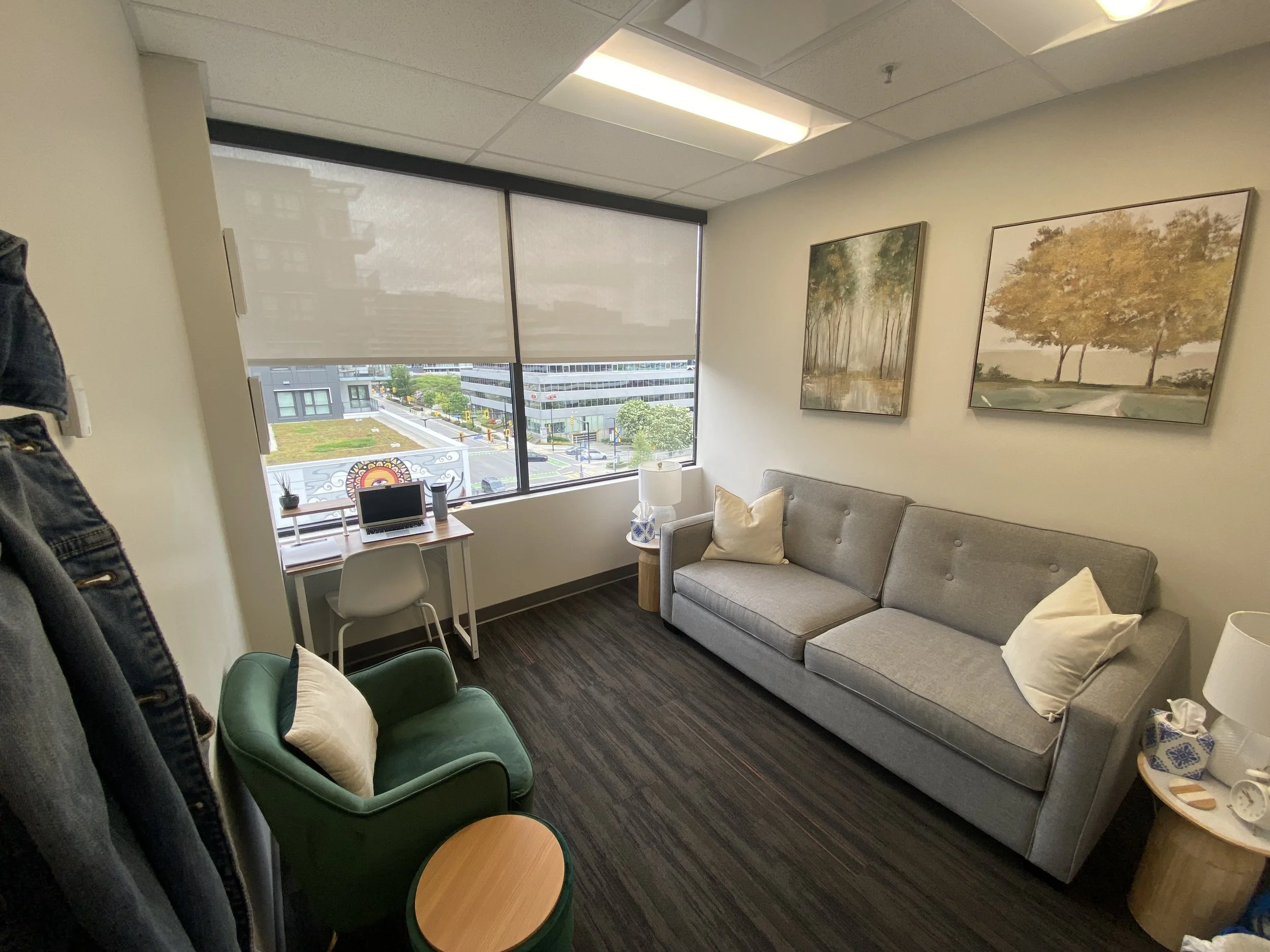 A modern office with a gray sofa, green armchair, side tables, lamps, and framed landscape paintings on the wall. Large windows with a view of a city street.