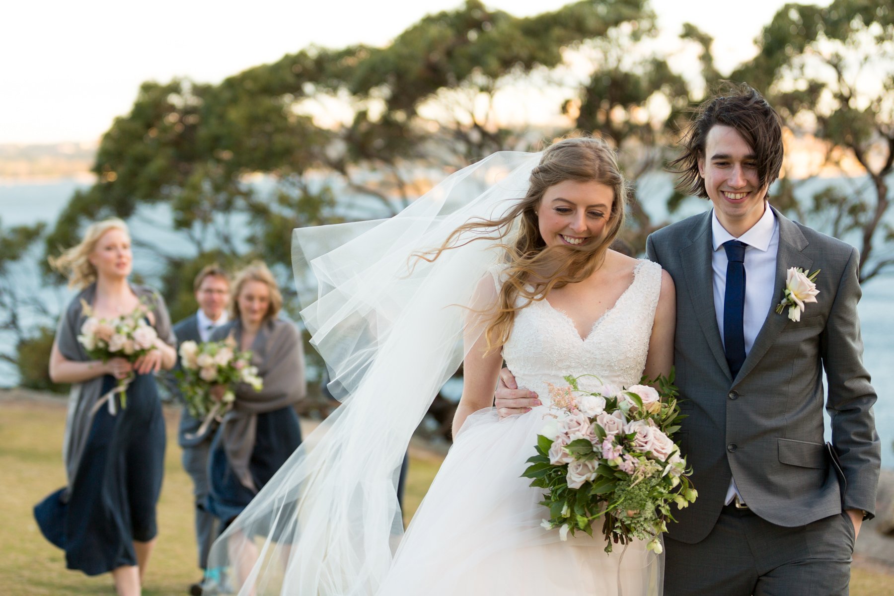 Bride and groom walking with bridesmaids and groomsman holding bouquets, outdoors wedding setting.
