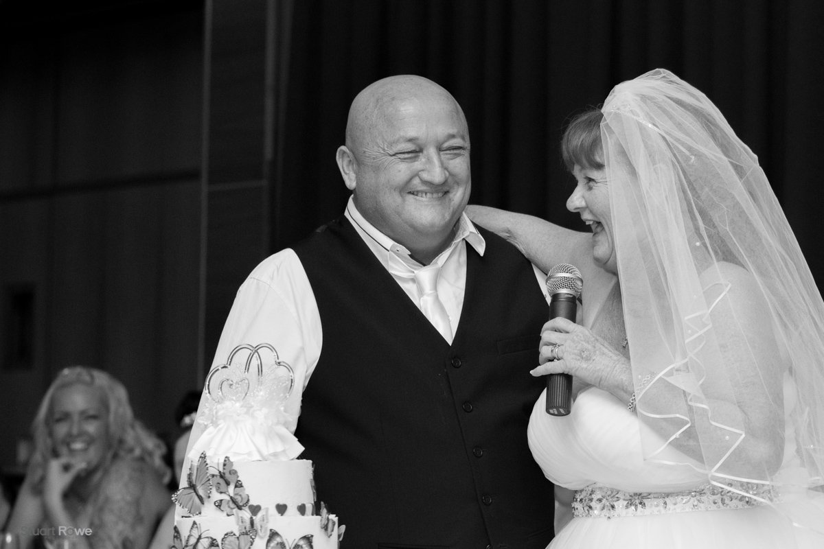 Bride and groom smiling during wedding speech with cake in foreground.