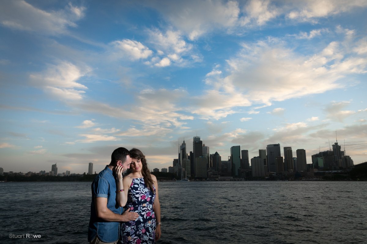 A couple standing by a body of water with a city skyline in the background under a cloudy sky.