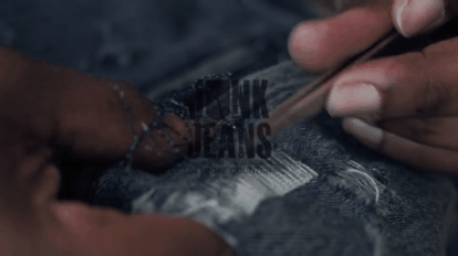 Close-up of hands using tools to repair or embroider denim jeans, with visible blue fabric and threads.