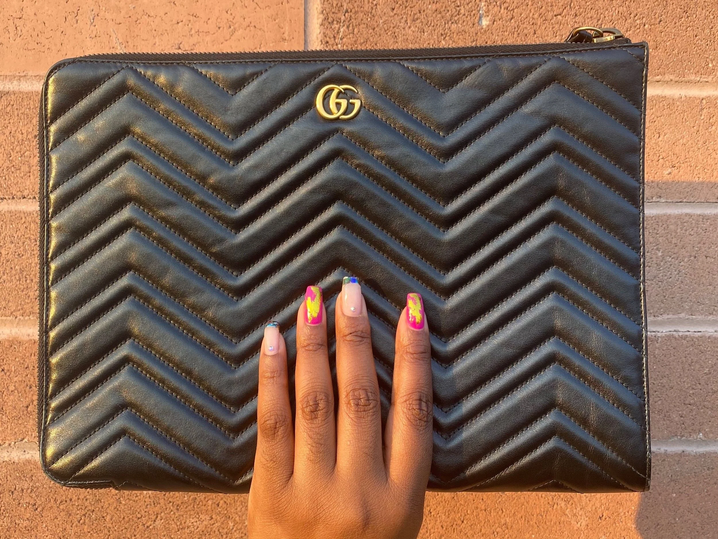 Black quilted clutch with zigzag pattern and gold logo, held by a hand with colorful nail art.