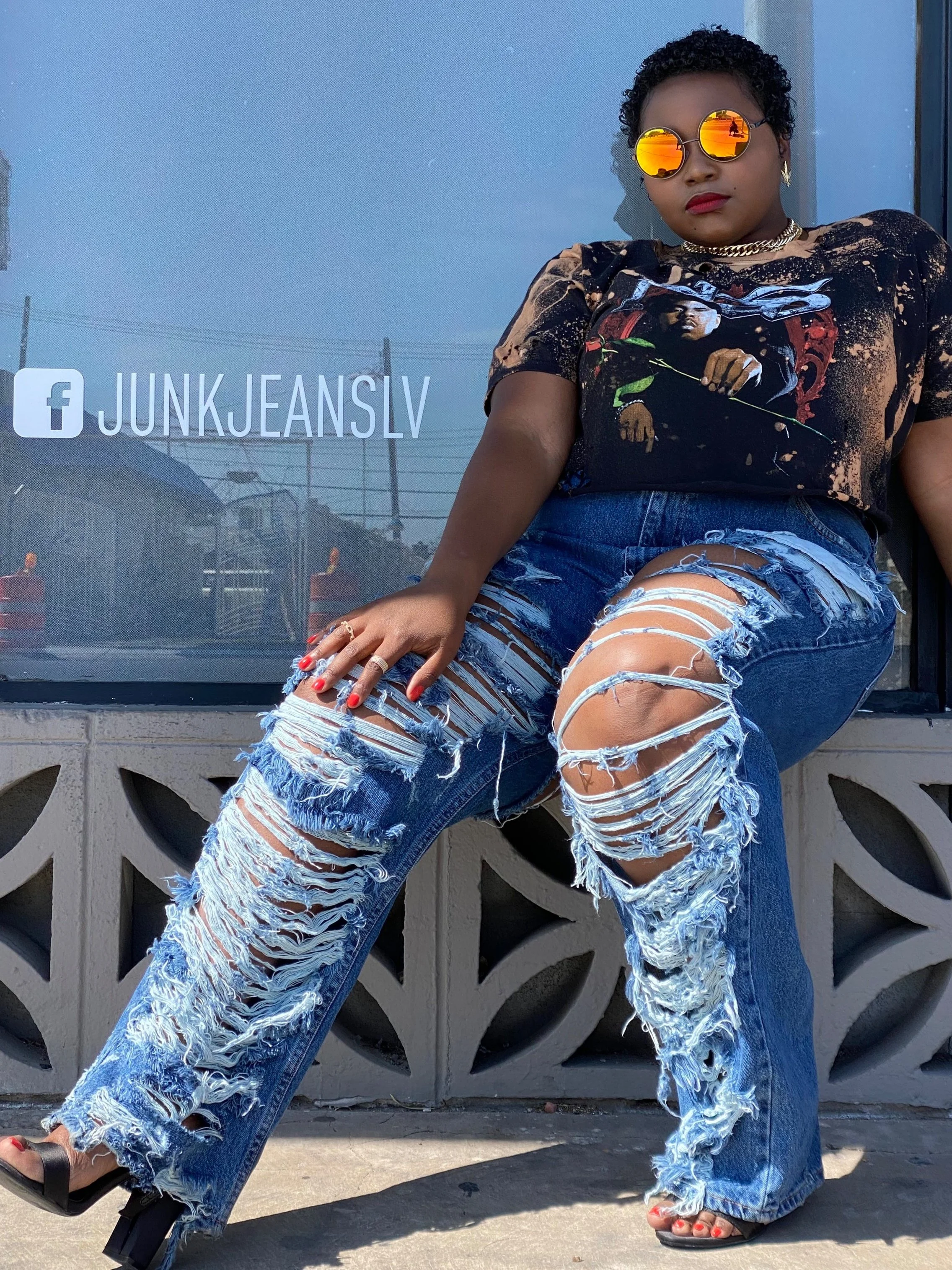 Person wearing heavily distressed blue jeans, band T-shirt, and reflective sunglasses, posing in front of a window with "JUNKJEANSLV" text on it.