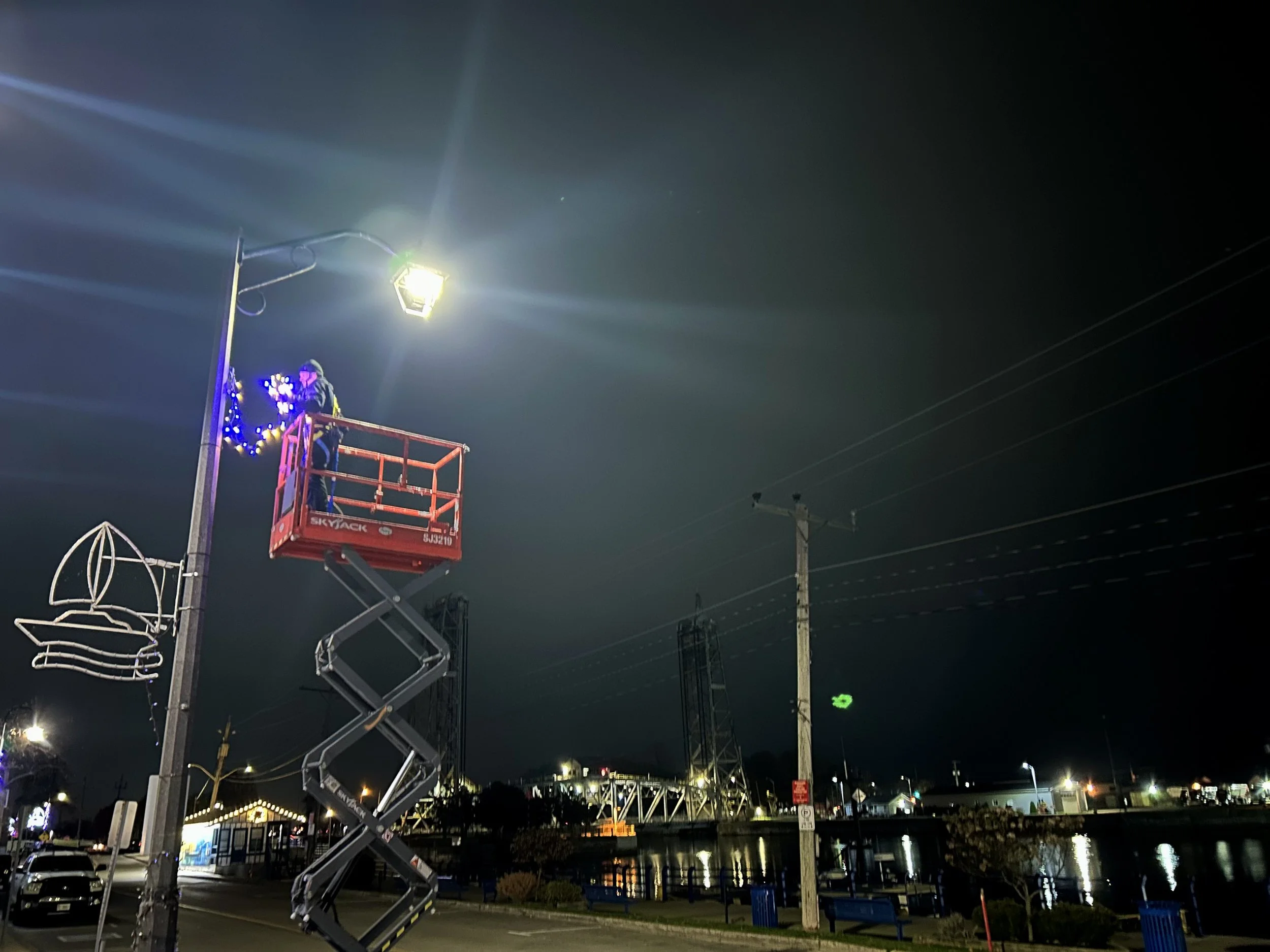 A worker operating a lift to attach decorative lights to a pole at night, with a dark sky and street lights in the background.