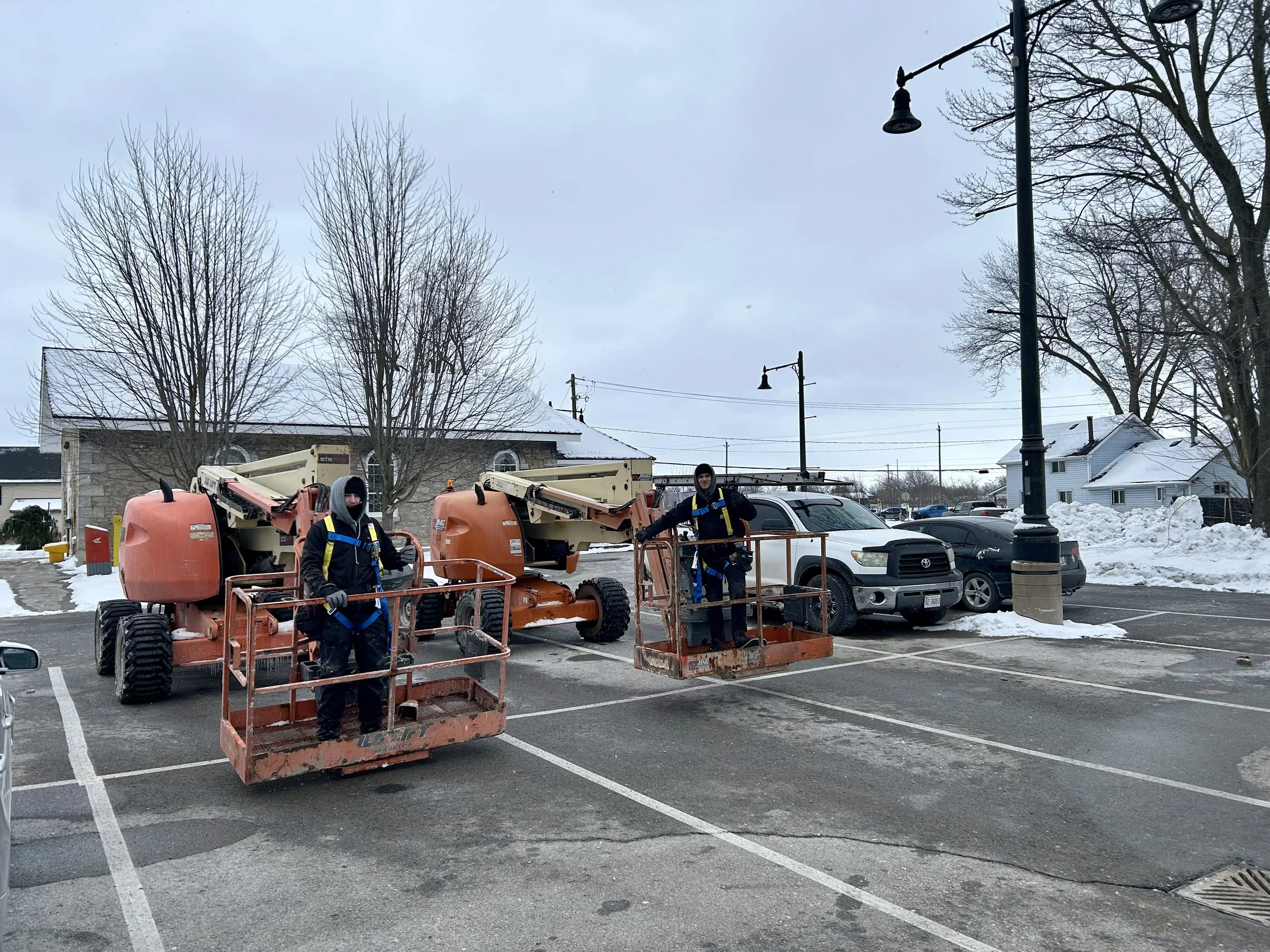 Two workers in winter clothing are standing on a cherry picker lift next to a parking lot with some snow. They are surrounded by parked cars, leafless trees, and streetlights, with cloudy skies overhead.