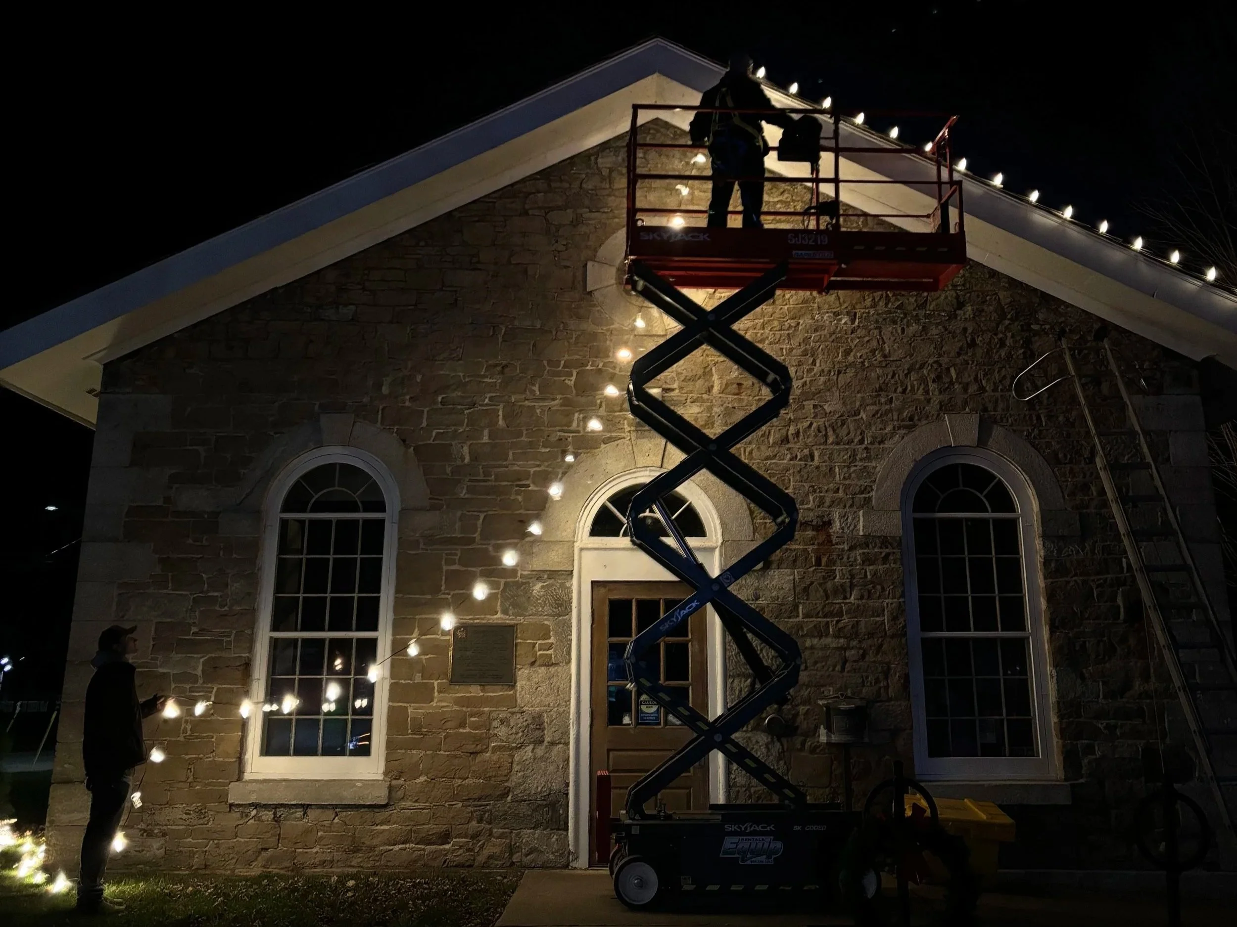 Night scene of two workers on a scissor lift installing string lights along the roofline of a stone building with arched windows.