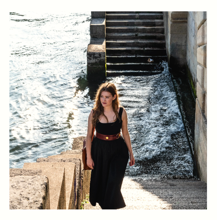 A woman in a black dress with a red belt walking up stone steps near water with a stone wall and stairs behind her.