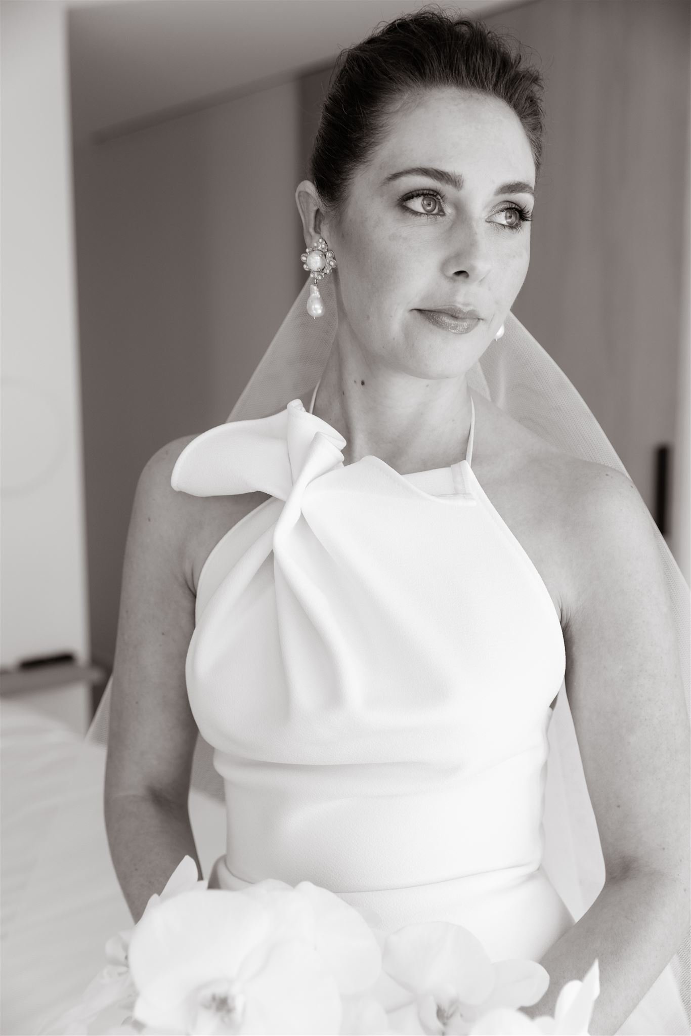 Bride in a white dress with floral detail and pearl earrings, holding white flowers, looking thoughtfully to the side.