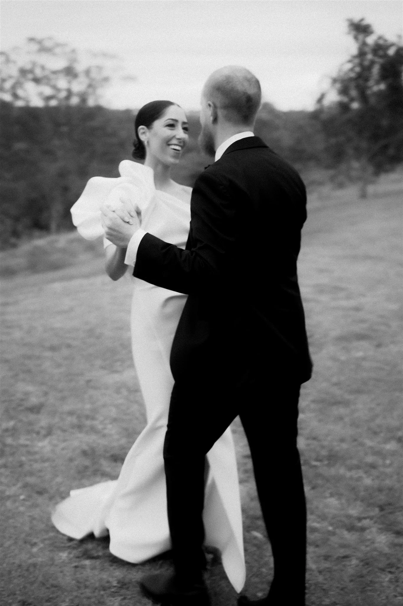 A couple dancing outdoors, the woman in a wedding dress and the man in a suit, smiling at each other.