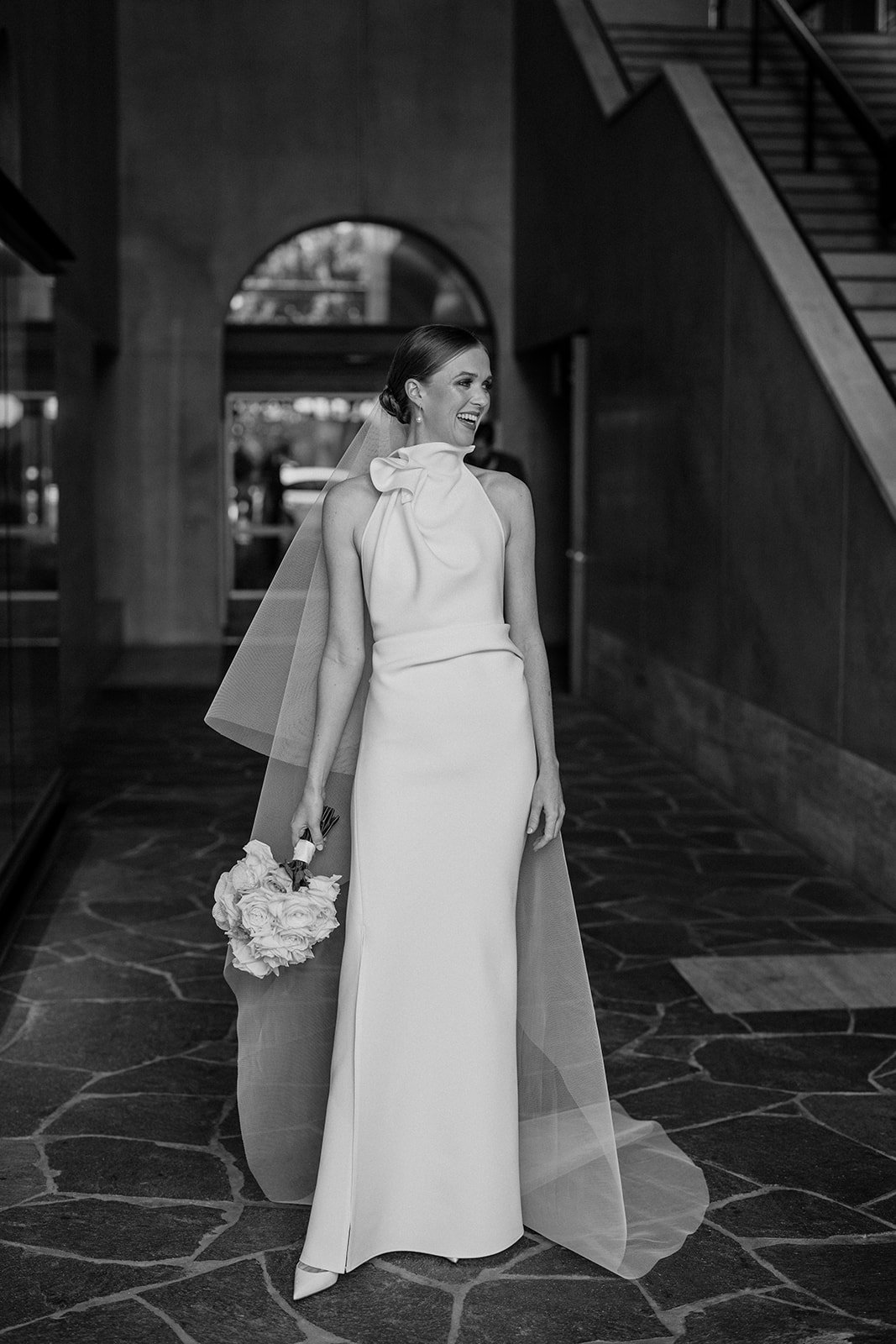 Bride in elegant wedding dress holding bouquet, standing in indoor setting with staircase.