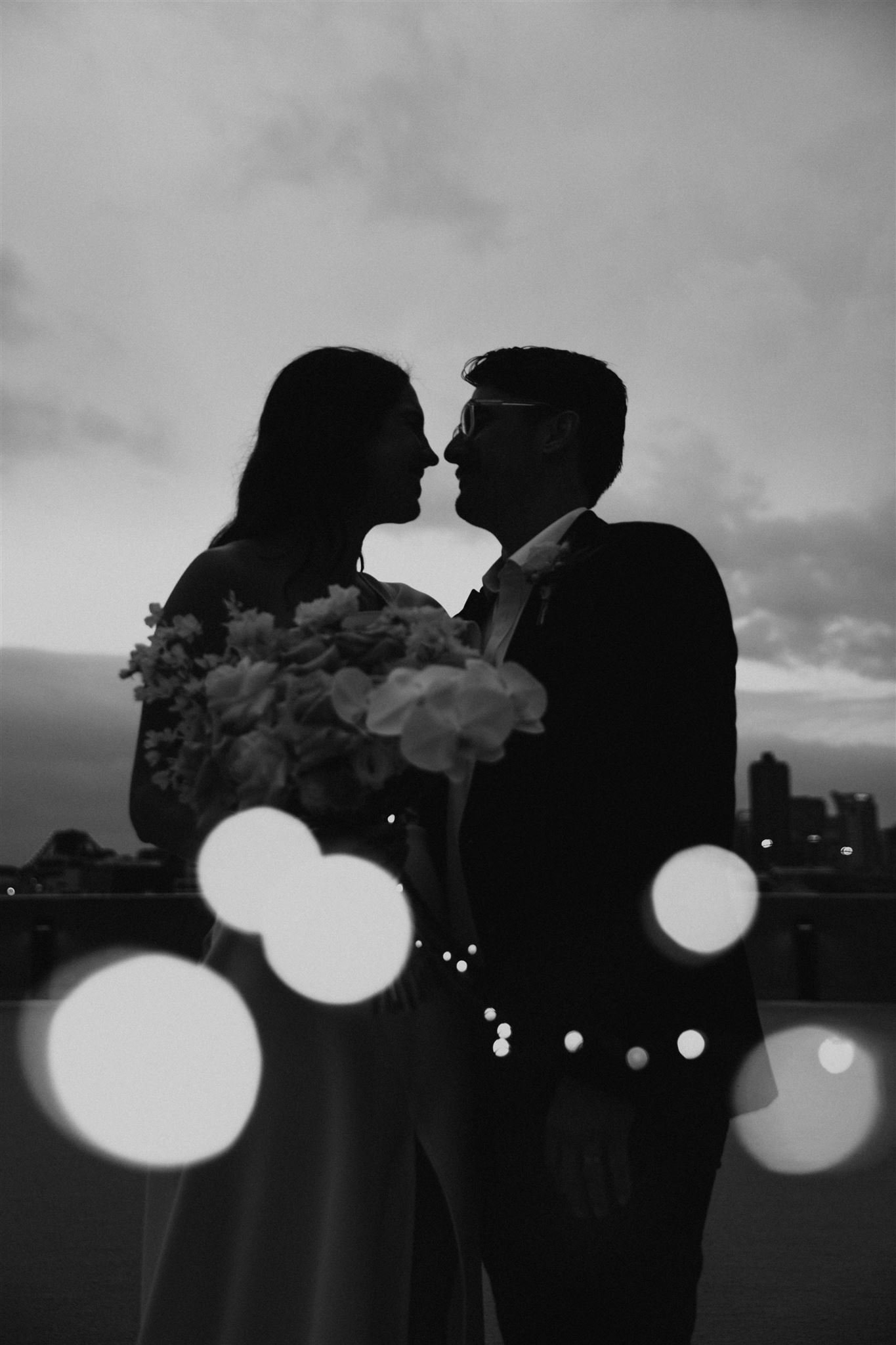 Silhouette of a couple embracing, holding a bouquet with city skyline at dusk and bokeh lights in foreground.