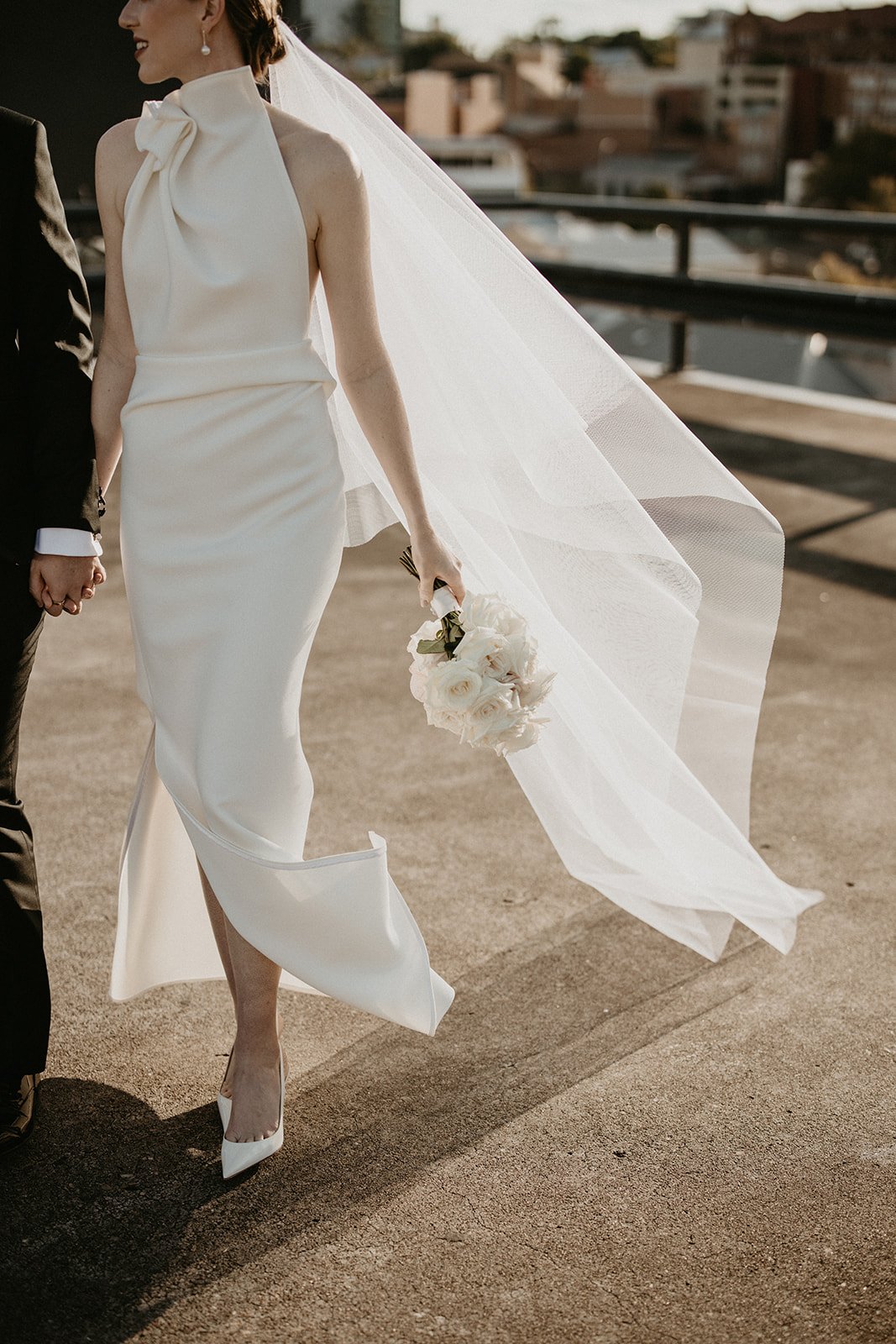 Bride in elegant white halter wedding dress with large bow and long veil, holding bouquet of white flowers, standing with groom in suit, on rooftop with urban background.