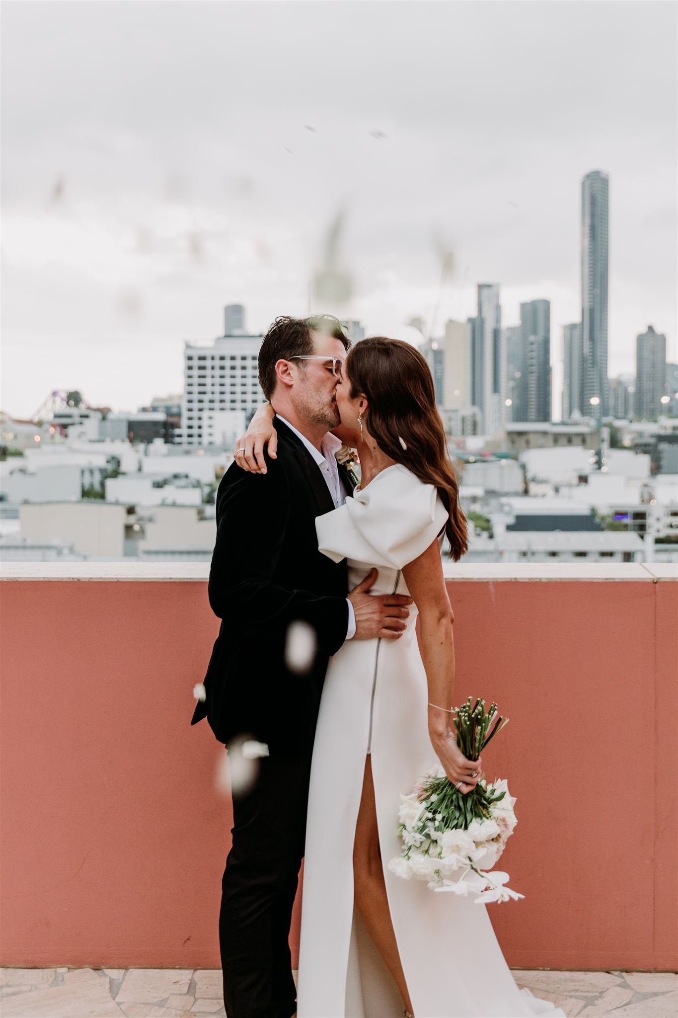 Bride and groom kissing on rooftop with city skyline background, bride holding bouquet of white flowers.