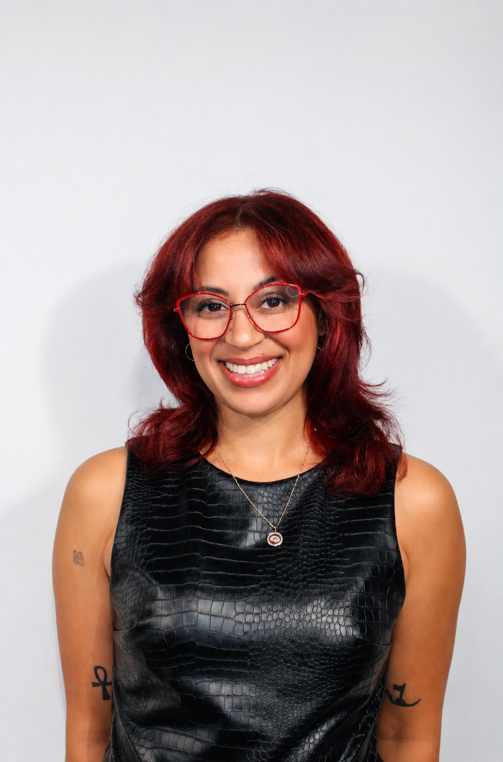 A woman with shoulder-length red hair wearing glasses, a black vinyl sleeveless top, and jewelry, smiling at the camera against a plain white background.