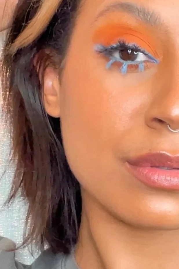 Close-up of a woman’s face showing her right eye, with orange eyeshadow, bright blue eyeliner, and matching blue eyelashes, along with part of her nose, lips, and brown hair.