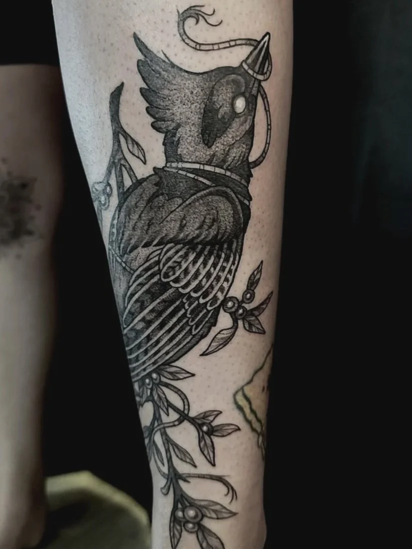 Black and gray tattoo of a bird with a rope, wing, and surrounded by leaves on a person's calf.