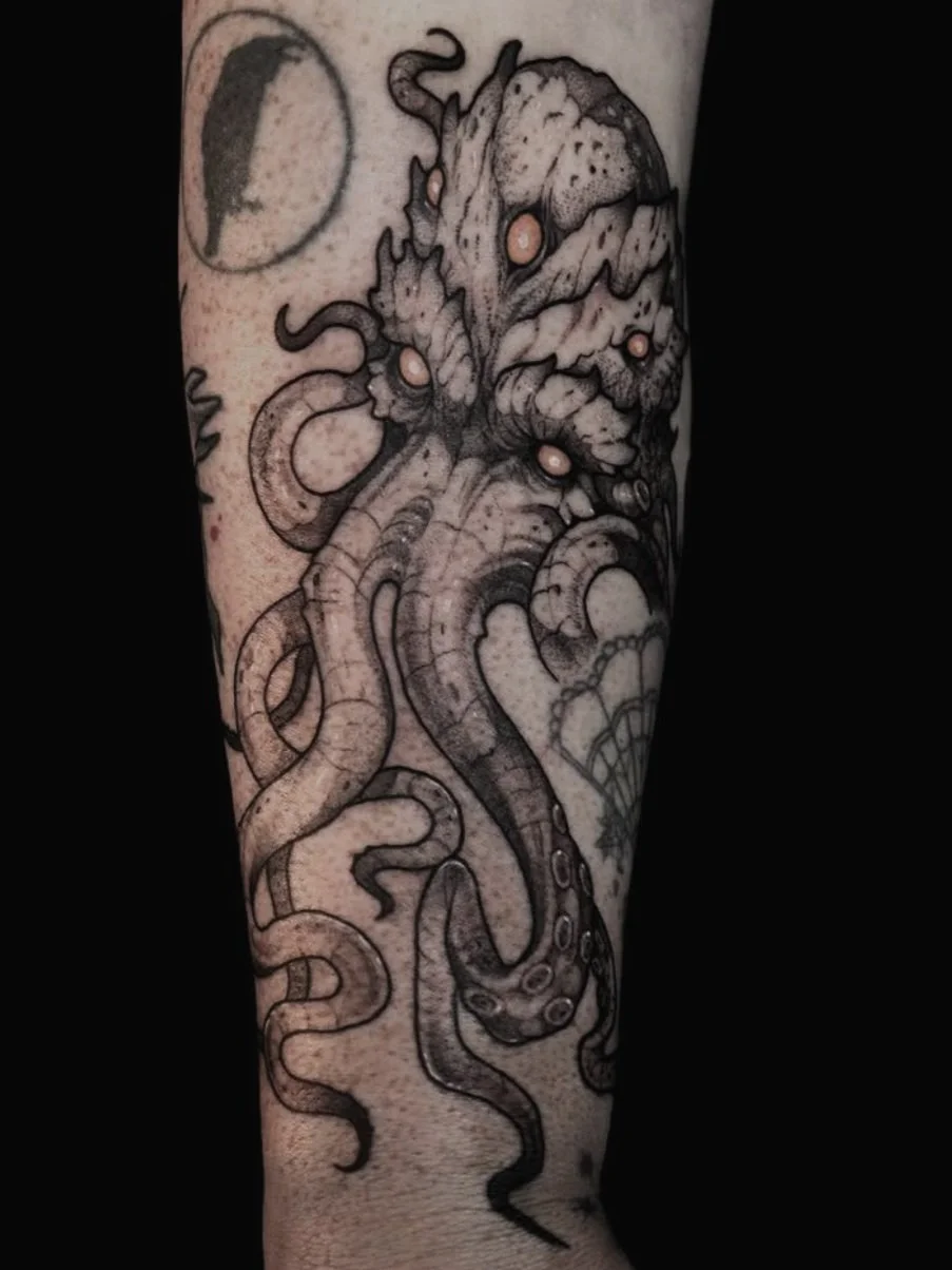 Black and gray tattoo of an cthulu-like octopus with glowing eyes on the forearm.