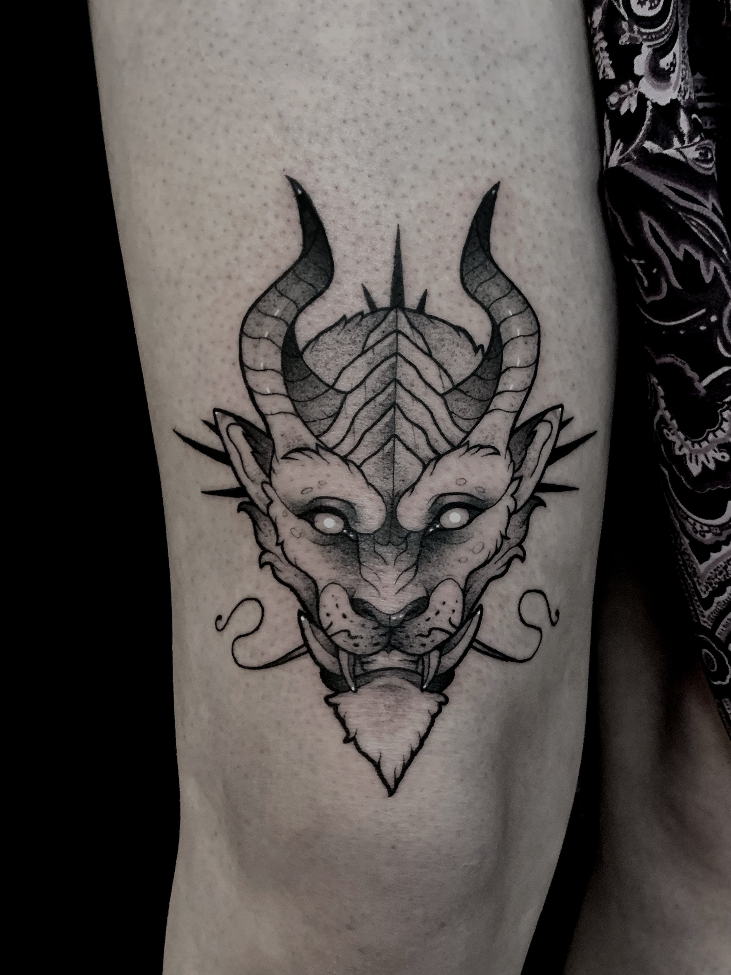 Tattoo of a stylized mythical creature with a feline face, horns, and decorative details on a person's arm.