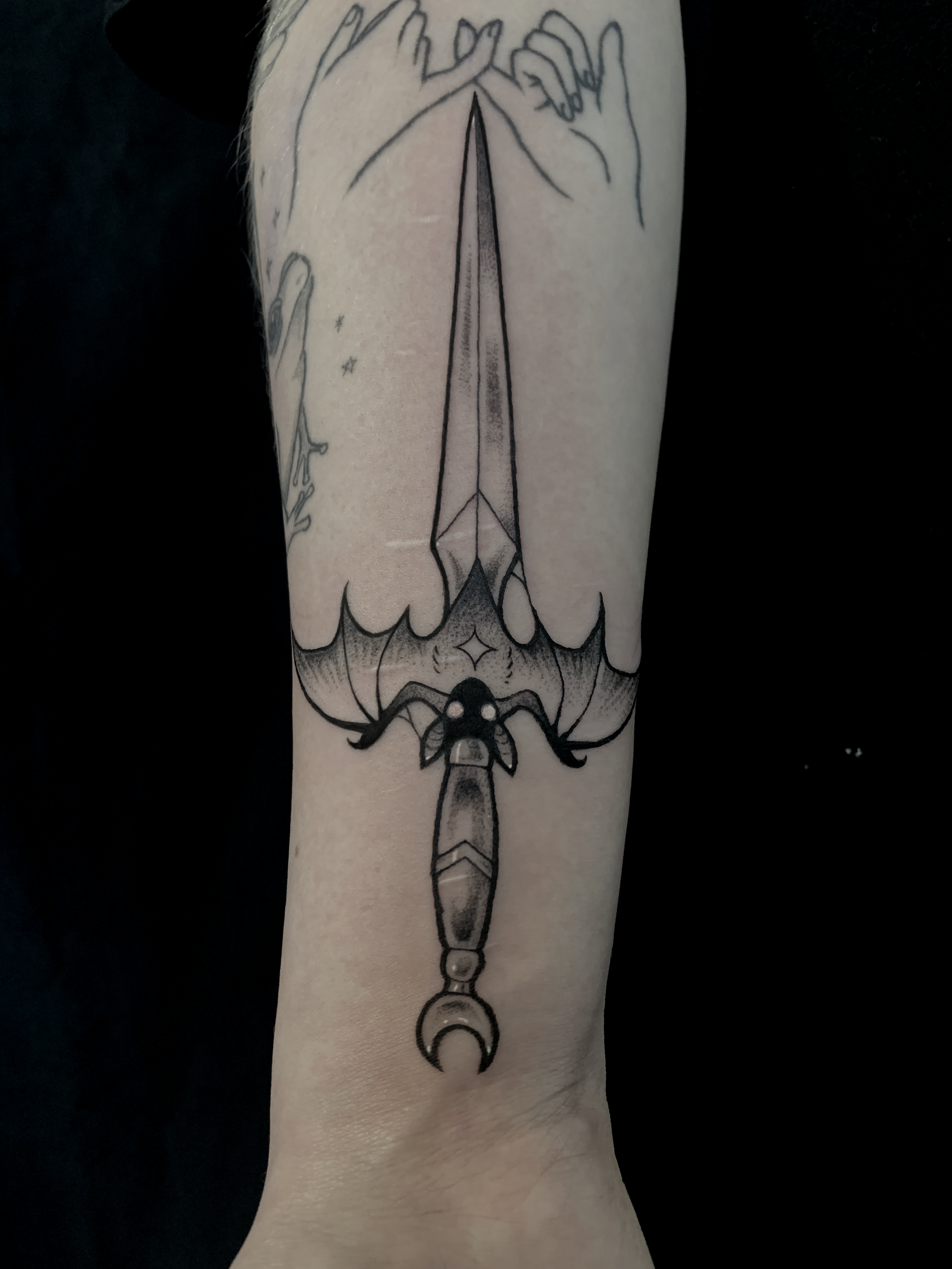 Tattoo of a dagger with an ornate hilt and a thin blade, surrounded by clouds, with a star and crescent moon in black ink on a person's arm.