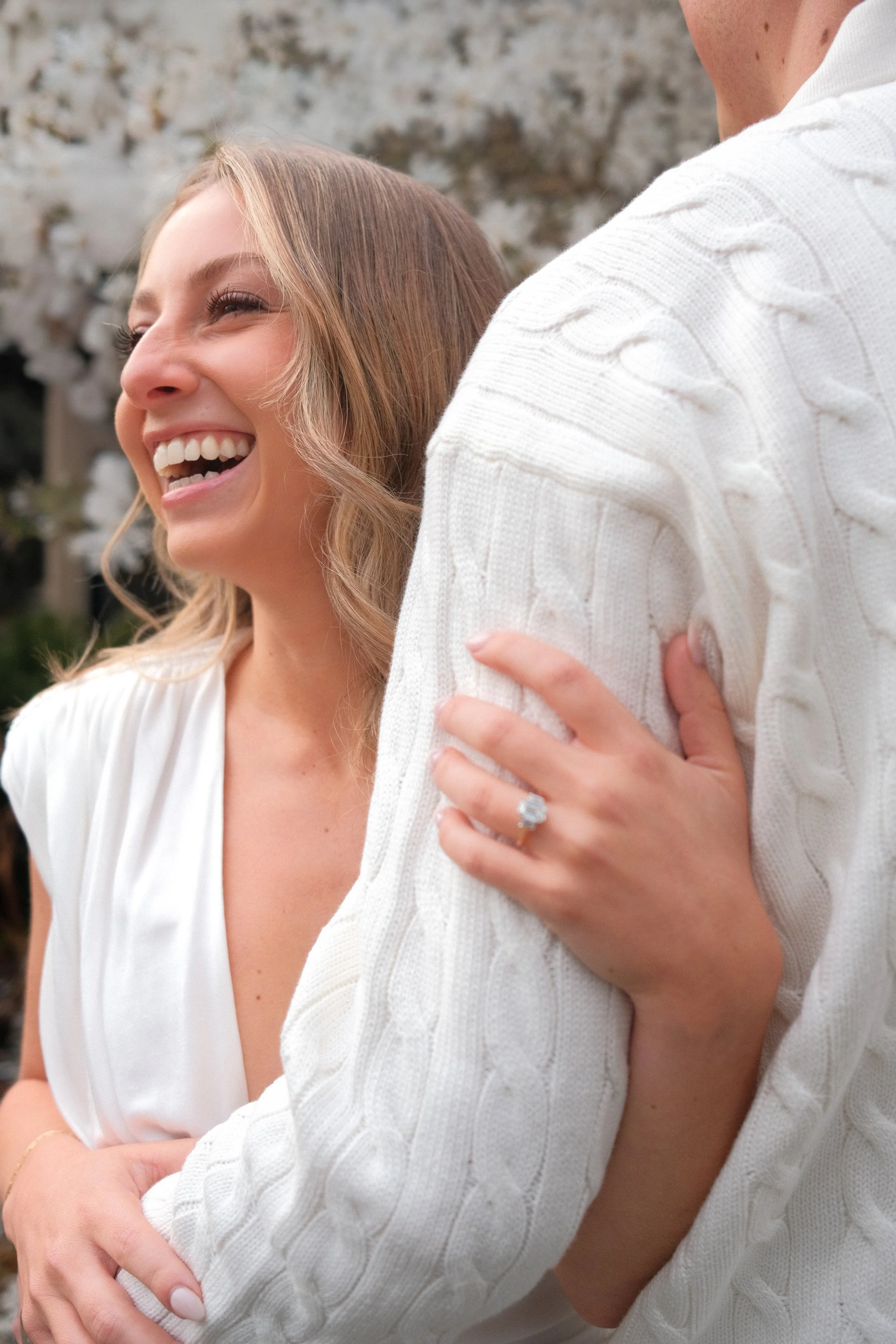 Close-up of a smiling woman with long hair and a man hugging her from the side, showing only part of his face and arm, both wearing white clothing. The woman wears an engagement ring.