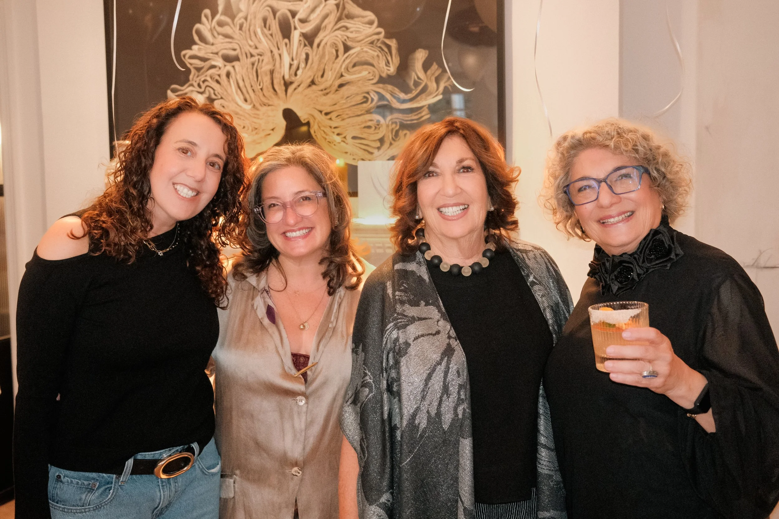 Four women smiling and posing together at a social gathering. One woman on the far right is holding a drink in a short glass.