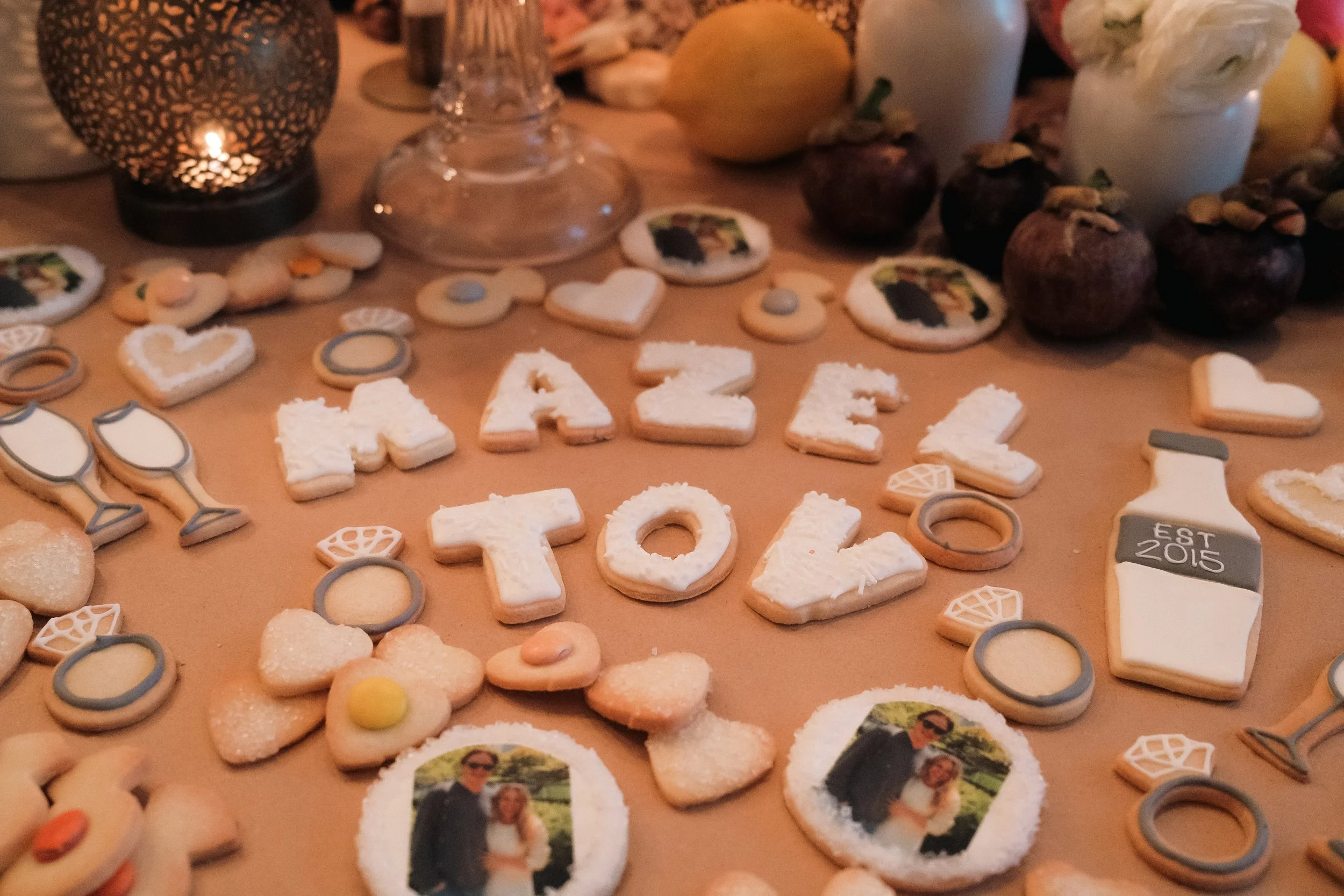 Decorative cookies, some with printed photo images, spelling out 'FAMILY' and 'TOGETHER,' with various other cookies and small candies, on a table with candles and fruits in the background.