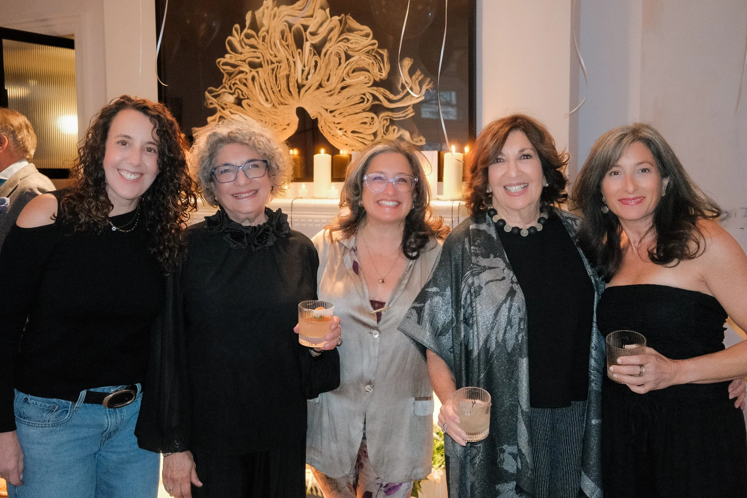 Five women standing together at a gathering, holding drinks, smiling, with a festive background of candles and an abstract art piece.