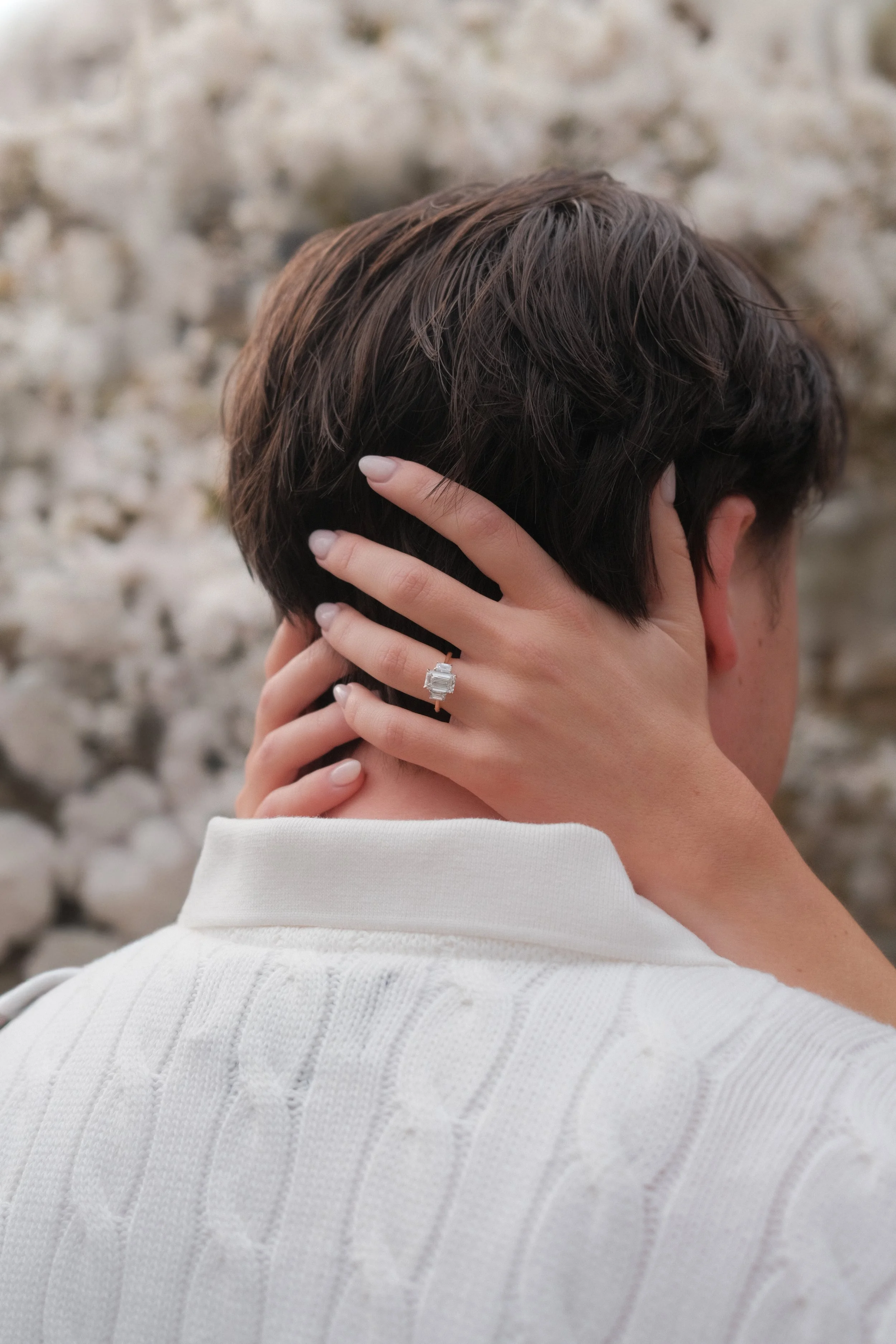 A person with short brown hair resting their head in their hands, wearing a white textured sweater, with a blurred stone wall background.