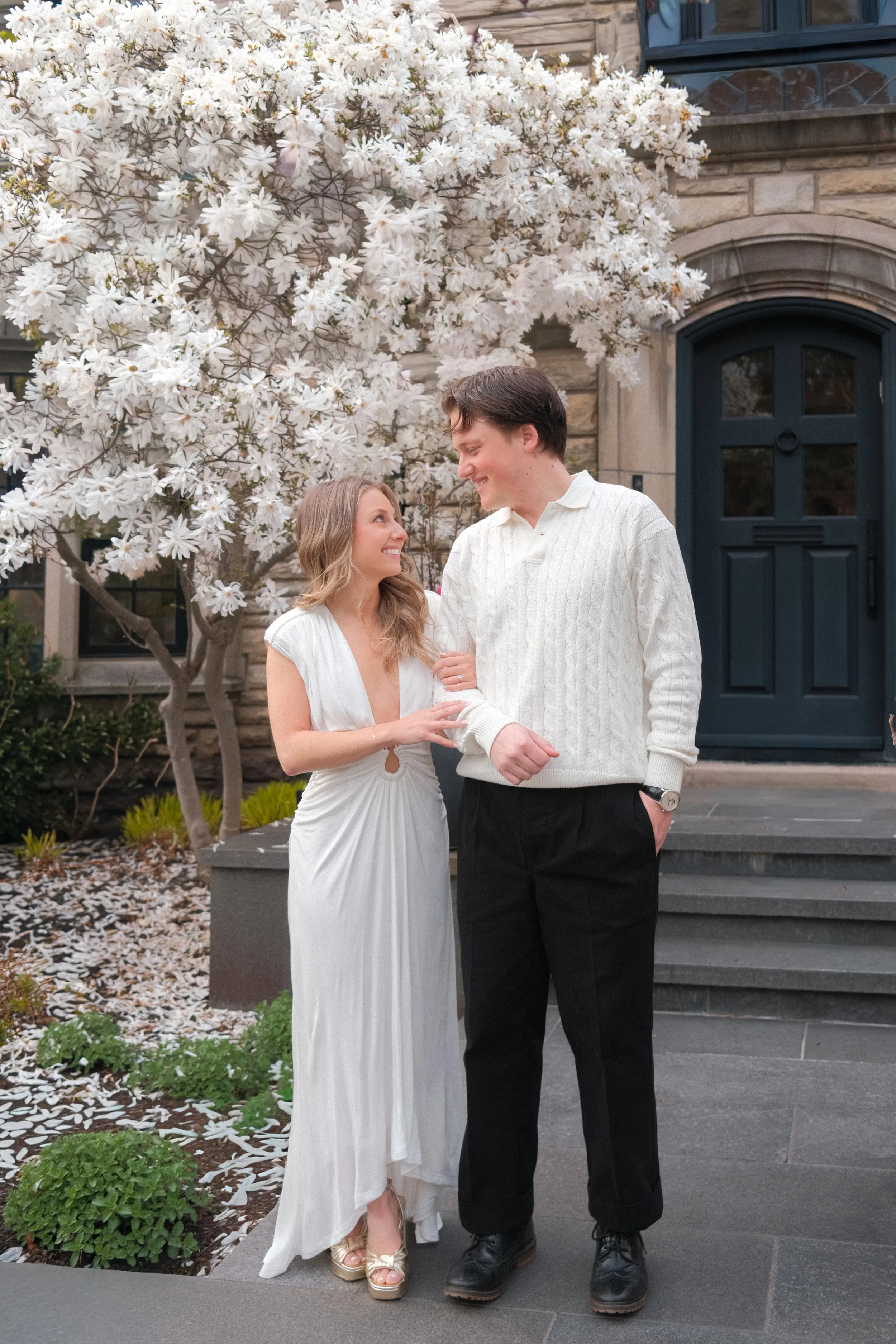 A smiling couple standing in front of a large white flowering tree outside a house with a stone facade and a blue door. The woman is wearing a white dress and sandals, and the man is dressed in a white sweater and black pants with boots.