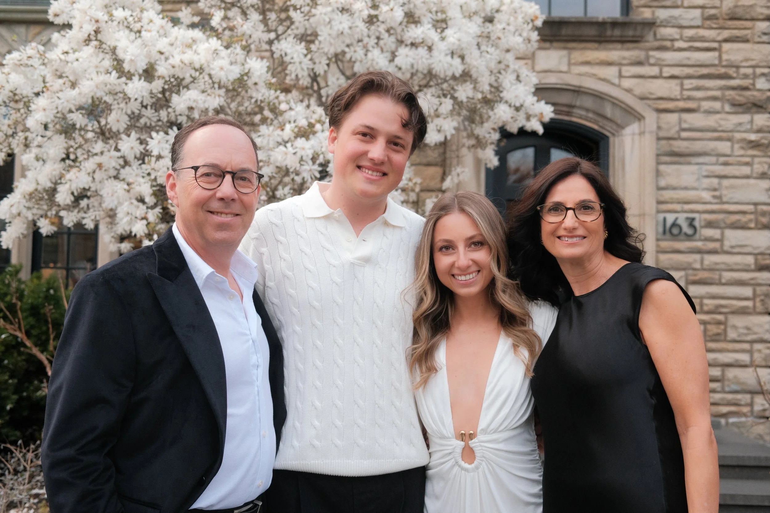 A family of five stands in front of a blooming white flower tree and a brick house, smiling at the camera.