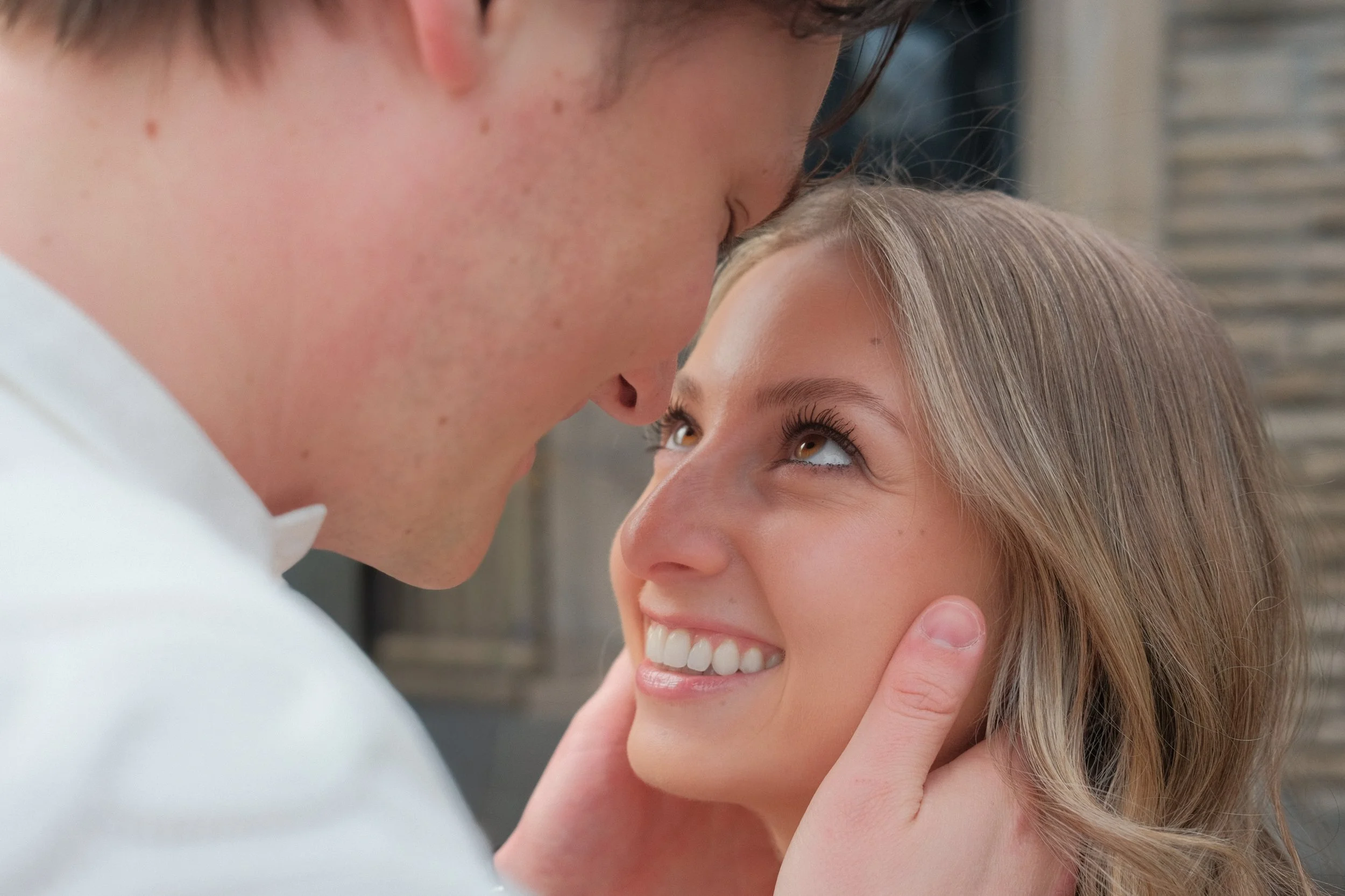 Close-up of a smiling young woman with blonde hair and a man with short brown hair leaning their foreheads and noses together.