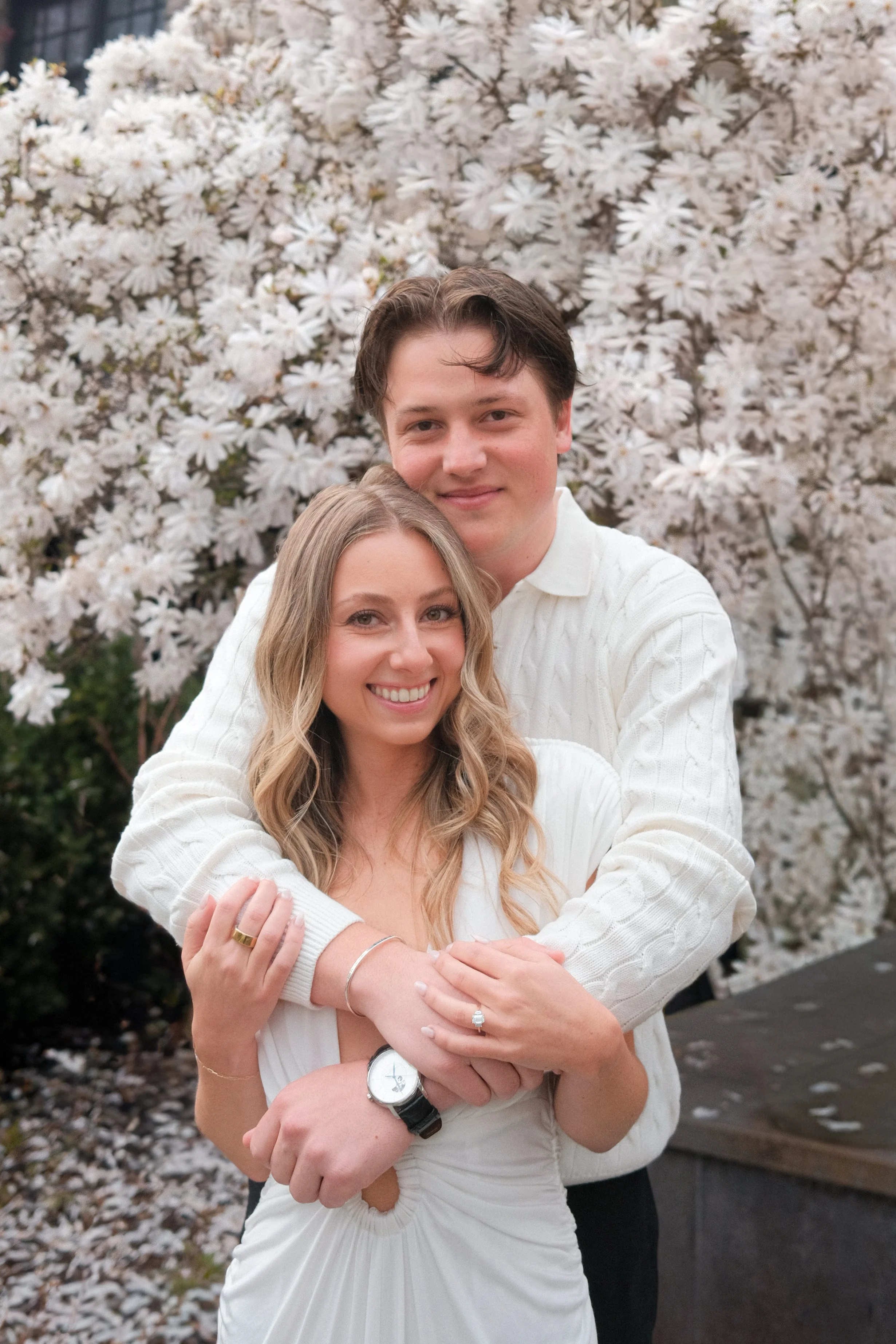 A couple happily embracing outdoors with blooming white flowers in the background.