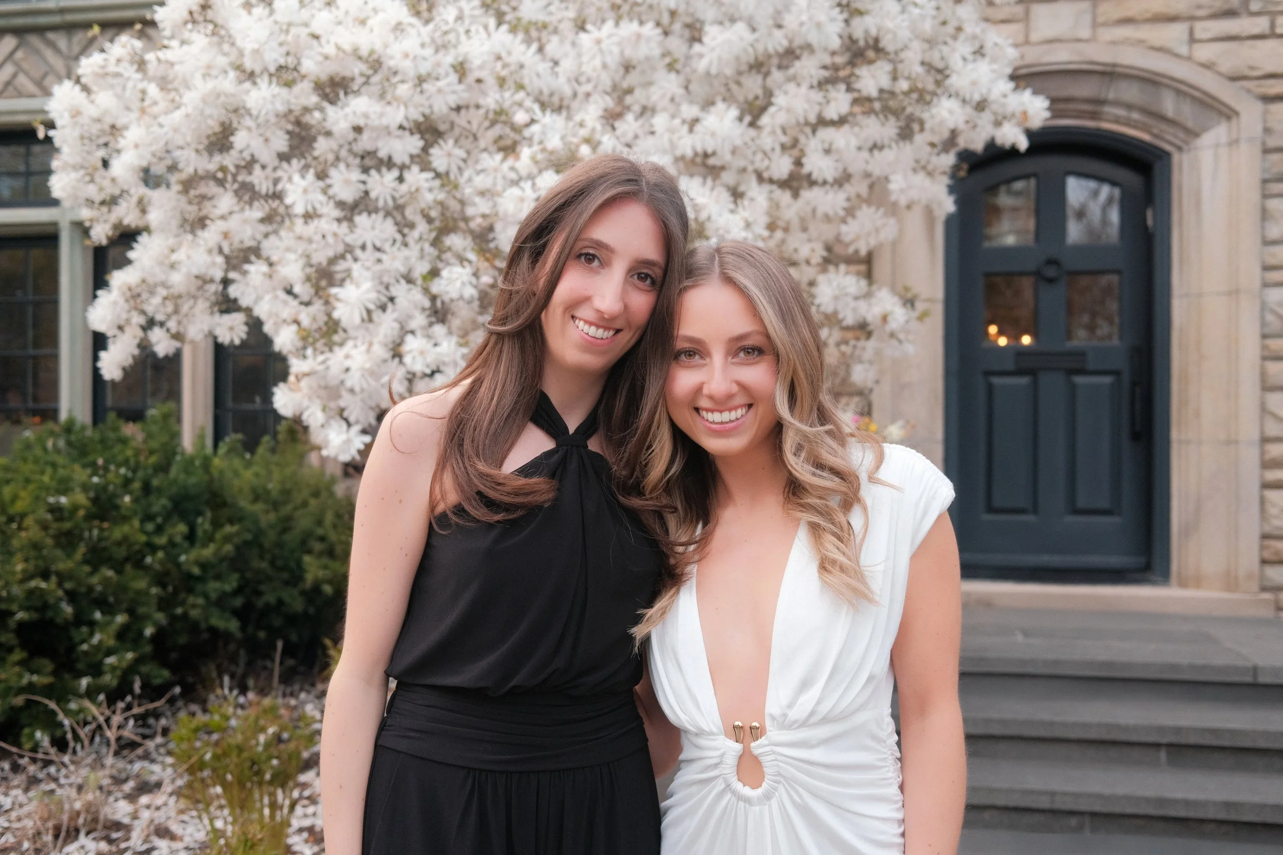 Two women smiling outside in front of a large white flowering tree, a dark blue door, and some bushes.